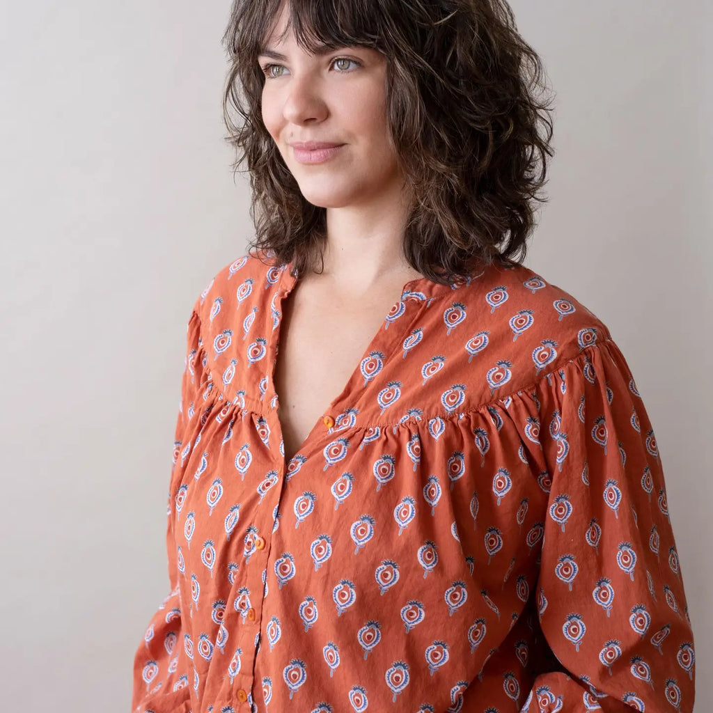 Woman wearing an orange patterned blouse and blue jeans sitting on a white surface.