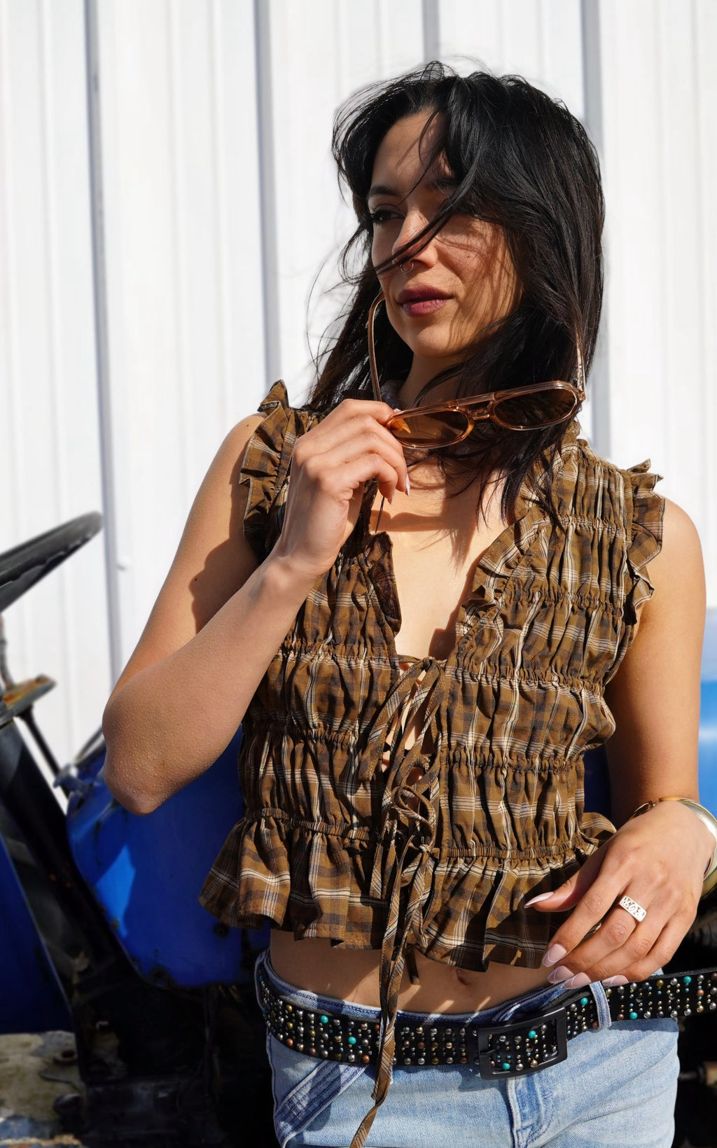 Woman wearing a fringed top and sunglasses with a white wall and metal object in the background