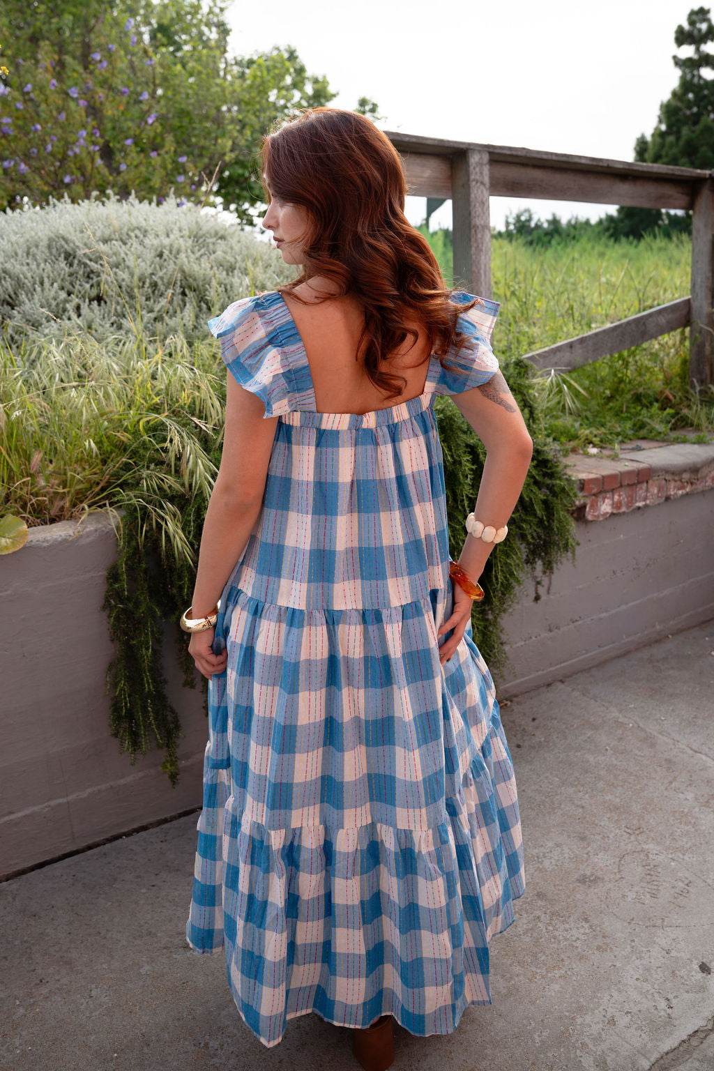 Woman wearing a blue and white checkered dress standing outdoors with greenery in the background