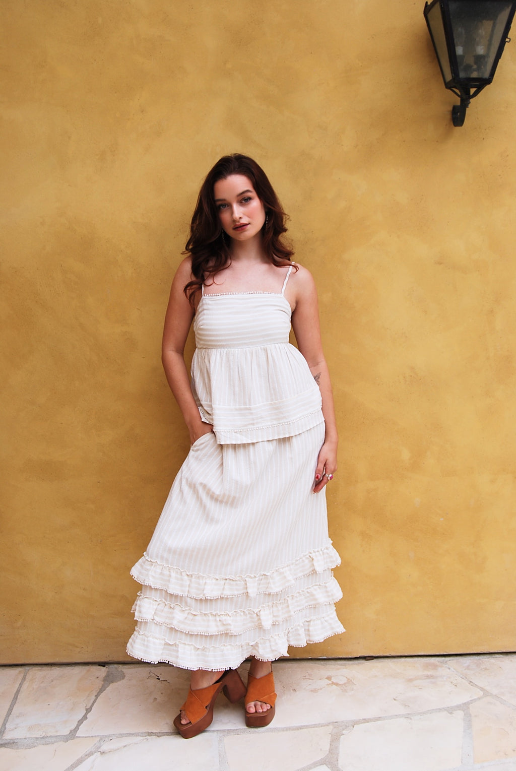 Woman in a white dress holding a straw bag against a yellow wall with greenery.