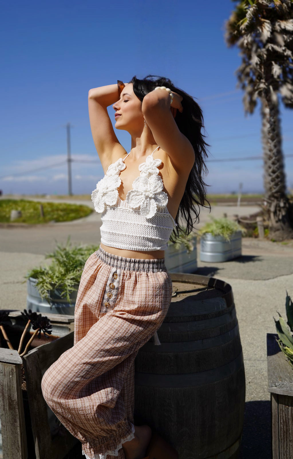 Person wearing brown checkered pants standing next to a wooden barrel with plants in the background.