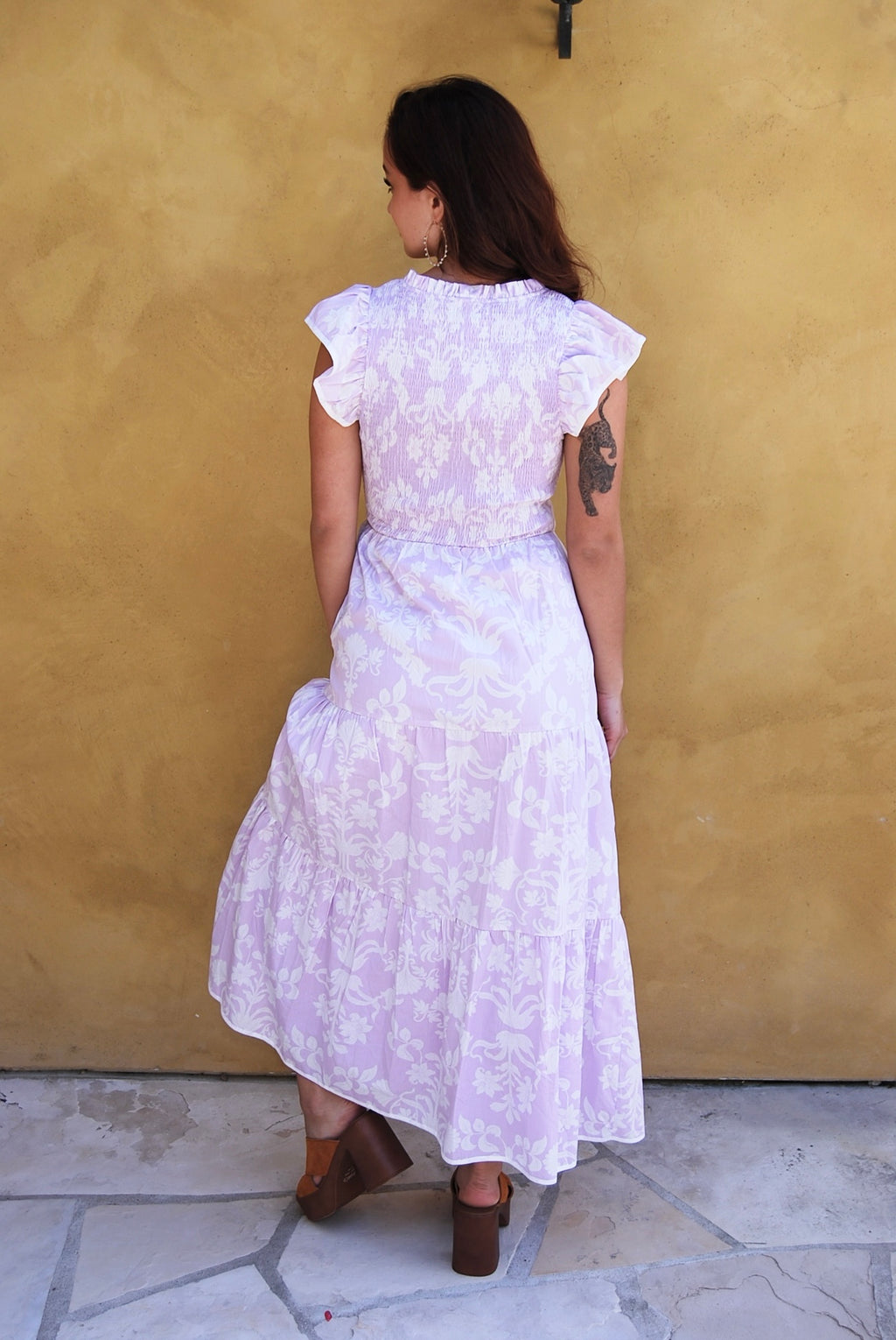 Woman in a purple floral dress standing in front of a yellow wall with flowers.