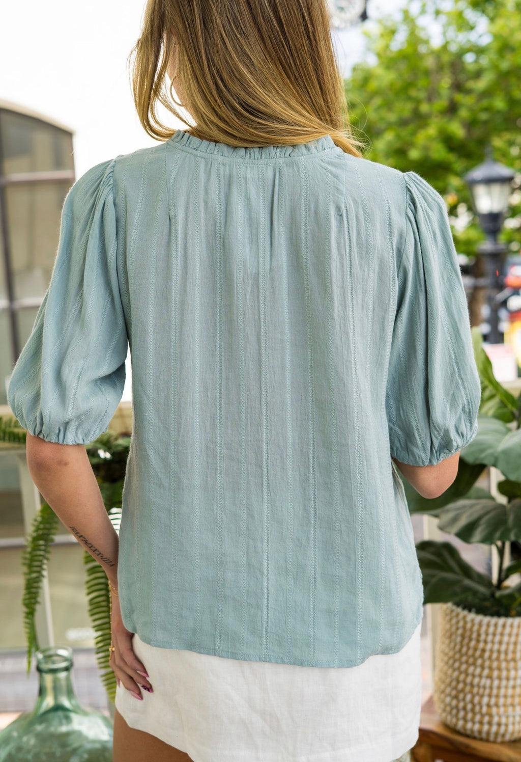 Woman wearing a light blue button-up shirt outdoors with greenery in the background