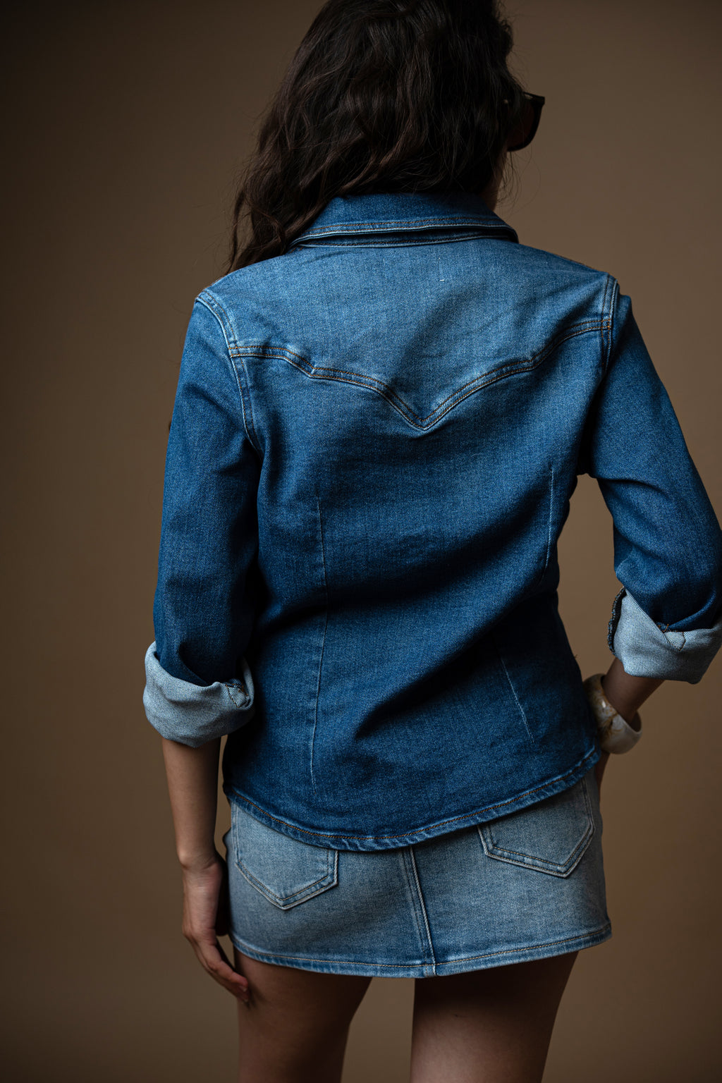 Woman wearing a denim outfit with sunglasses and a brown belt against a brown background
