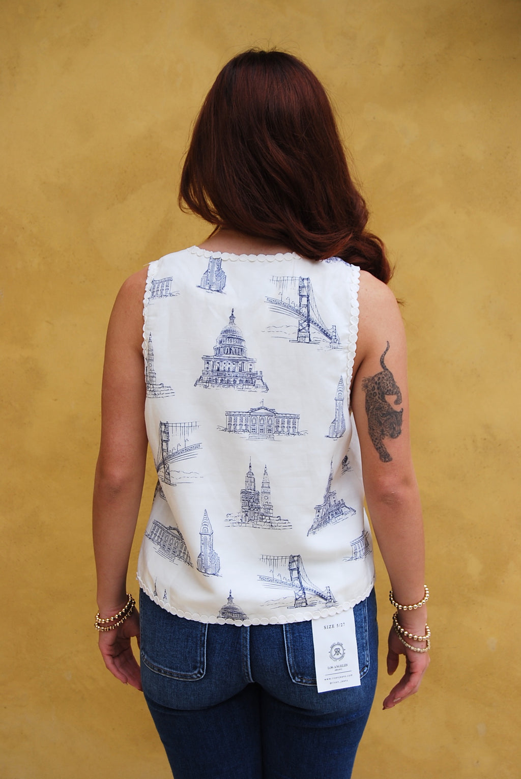 Woman wearing a patterned vest and blue jeans against a yellow wall